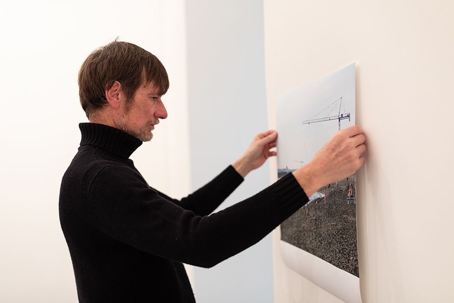 Color photograph of a light skinned man with brown hair wearing a black turtleneck; he is hanging an unframed photograph on a white gallery wall