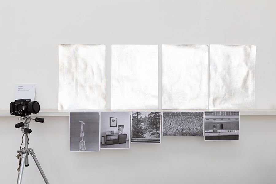 Color photograph of a studio wall with four large sheets of wrinkled paper above four black and white 8x10 photos; to the left is a camera mounted in a tripod
