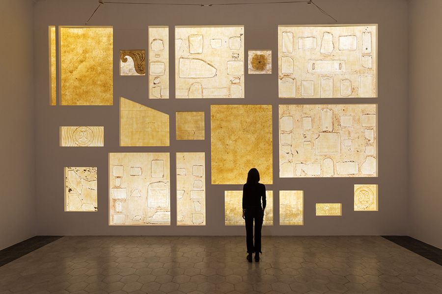 Color photograph of a woman standing in an indoor gallery in front of an art installation comprising pieces of latex of various sizes bearing the imprint of ancient inscriptions