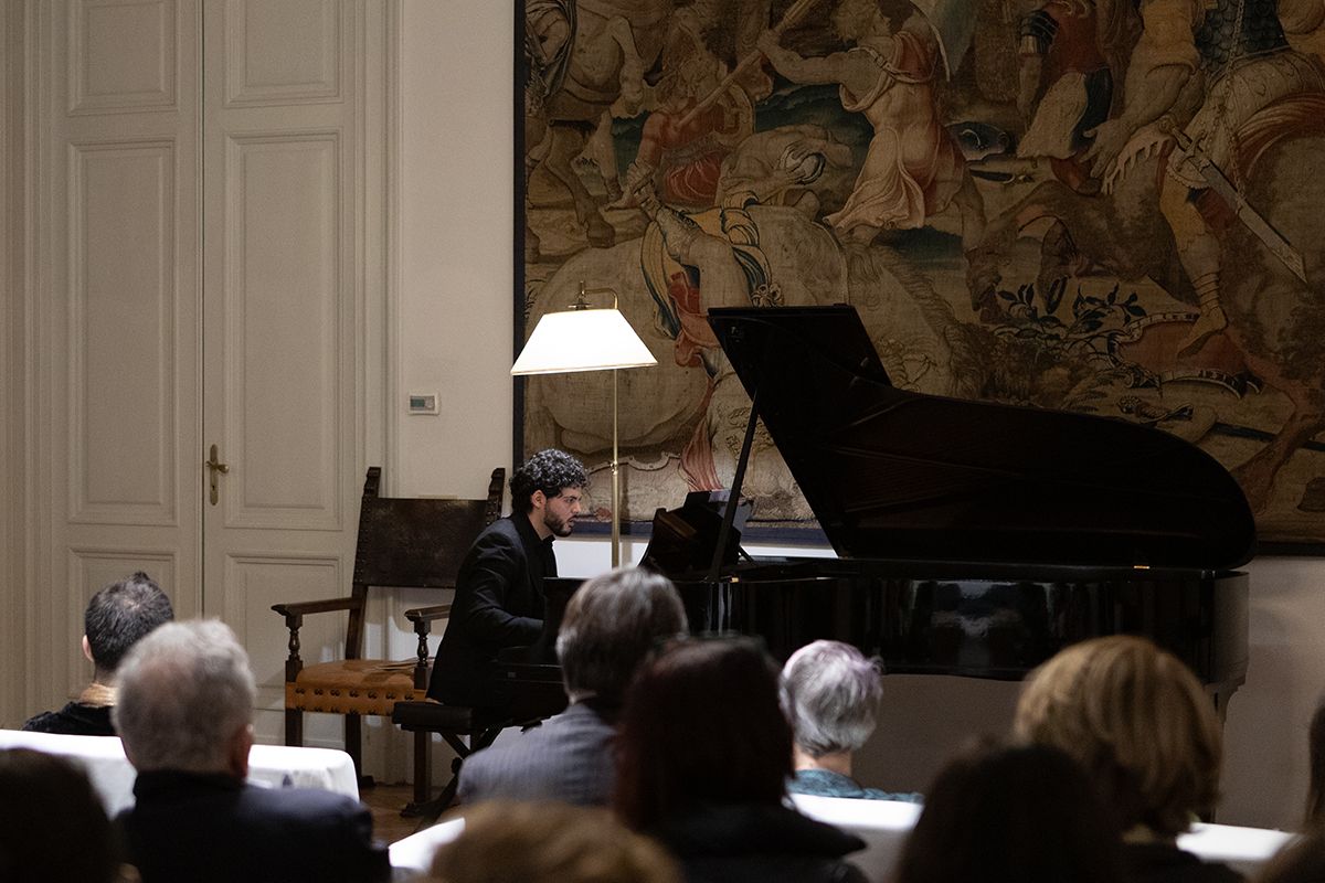 Color photo of a man playing piano in front of a tapestry and a seated audience