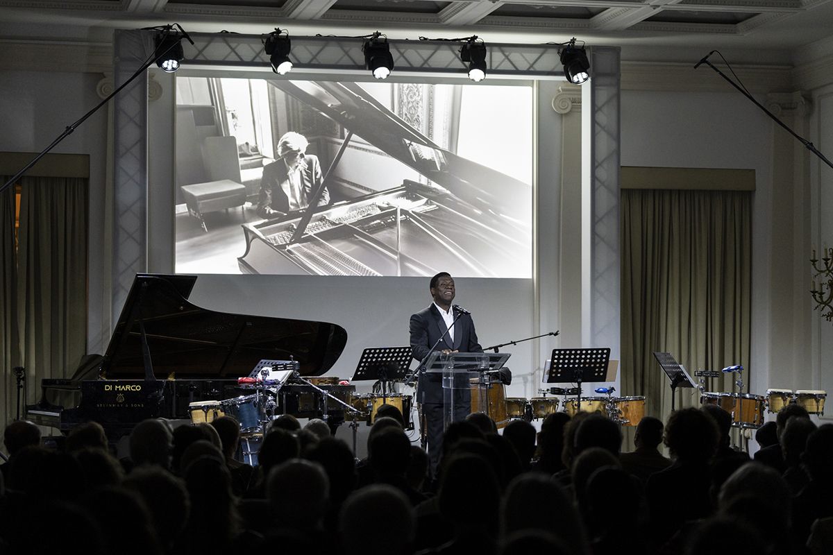 A dark skinned man stands at a podium overlooking an audience with a photograph of a pianist projected on the wall; behind him is a piano, several sheet music stands, and various percussion instruments
