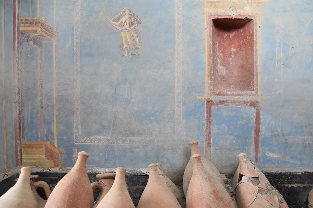 Color photo of an excavated room in Pompeii with blue walls and clay amphorae in the foreground