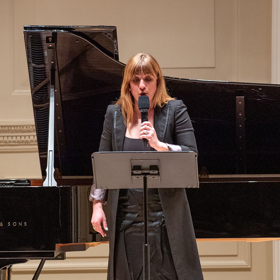 Color photo of a light skinned woman singing onstage holding a microphone, with a grand piano behind her