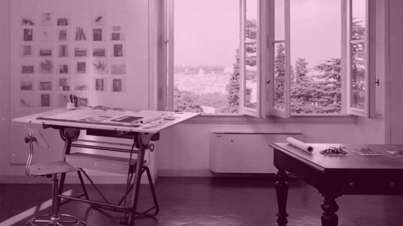 Photograph of an artist's studio showing a work desk, stool, wooden table, and open windows