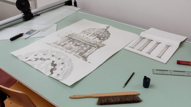 Color photograph of a desktop with a large pencil drawing of a frontal view of Bramante's Tempietto, a small circular temple; also on the desktop are a pencil, pencil sharpener, another drawing  of four columns, a sweeping brush, and a ruler