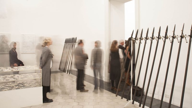 Installation view of Terry Adkins's work "Meteor Shower" at AAR in 2015, showing people milling about a room with pikes (pole weapons) leaning on the wall in a row
