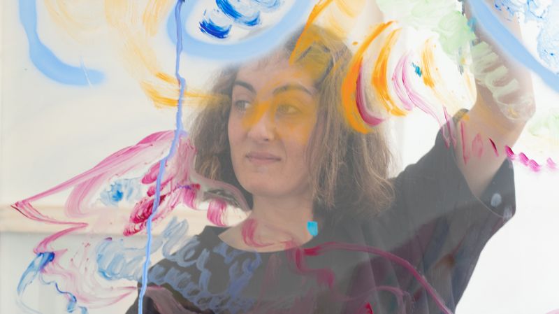 Color photograph of the head and shoulders of a light skinned woman with brown hair; she holds a translucent sheet in front of her that is covered with colorful swirls