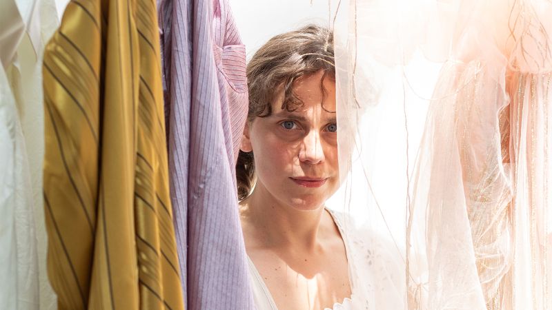 Photograph of the head and shoulders of a light skinned woman standing among hanging fabrics of many colors