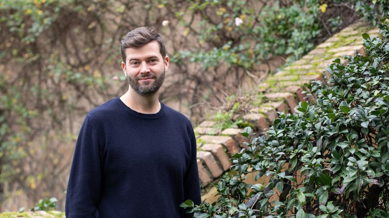 Color photograph of a light skinned man with a beard wearing a black sweatshirt and looking at the camera; he stands in a garden with a brick stairwall behind him