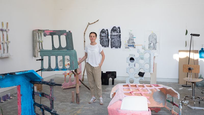 Color photograph of a light skinned woman wearing a white t-shirt and khaki pants; she stands in an artist's studio among many works of her sculpture