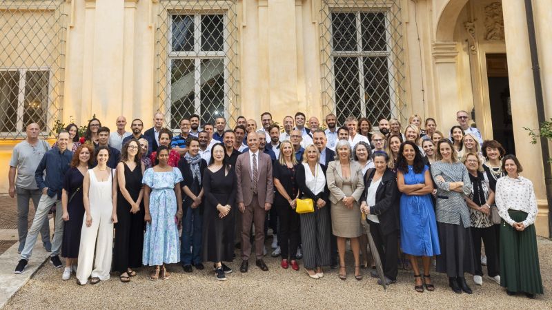 Color photograph of several dozen people standing in rows as they pose in front of a Roman villa