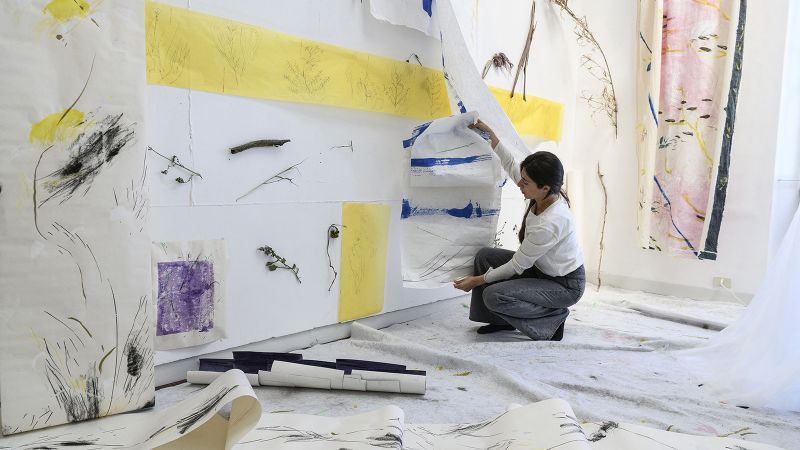 Color photograph of a light skinned browb haired woman crouching in a light filled artist's studio, with drawnings and paintings hanging on the wall and resting on the floor