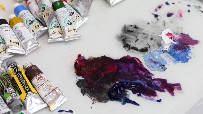 Color photograph of a tabletop in an artist's studio showing tubes of paint as well as paint from those tubes on a white sheet on the table
