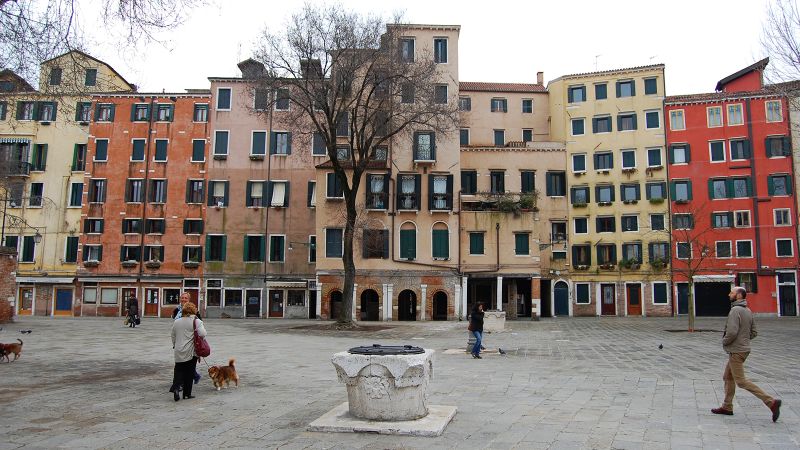Color photograph of Campo di Ghetto Nuovo in Venice, featuring an open plaza with people and dogwalkers in the foreground, and six and seven story residential buildings in the background, each painted in shades of red, tan, and yellow
