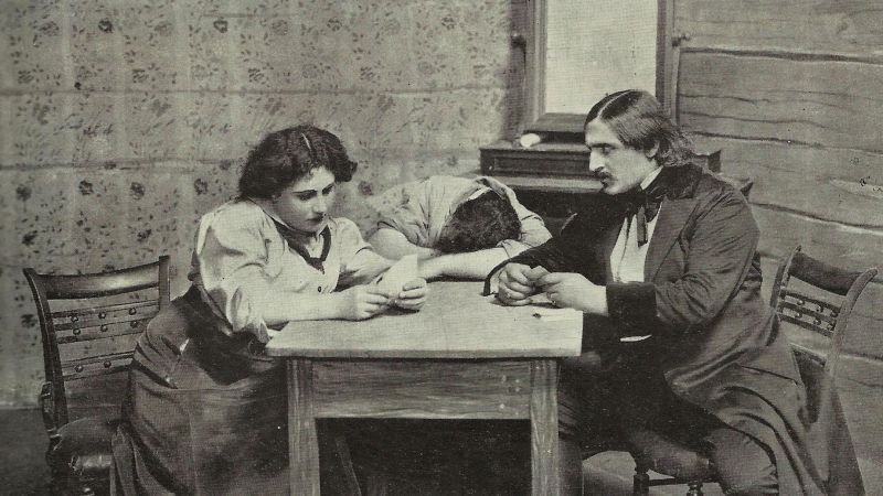 Black and white photograph from 1910 of a stage play depicting a woman and two men wearing early 20th century clothes seated at a table; one man has his head on the table sleeping