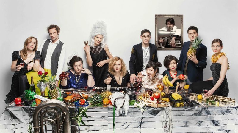 Color photo of ten people seated or standing around a long dinner table topped with fruit and musical instruments
