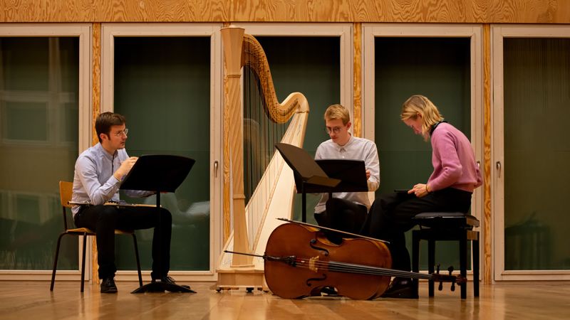 Color photo of three muscians, their instruments, and music stands in a room