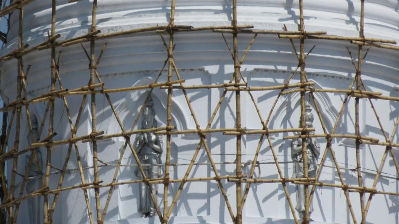 Color photograph of a bamboo or wooden scaffolding covering the Hatharas Kotuwa (tee cube) of a stupa; sculptures of three figures with their palms facing each other as if in prayer
