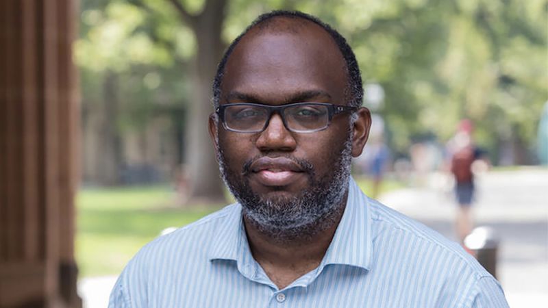 A man with glasses and a blue shirt, trees and park-like setting in the background