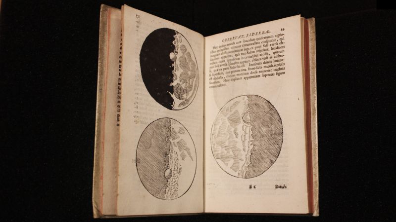 Color photograph of an open book against a black background; the book is open to pages with drawings of the moon