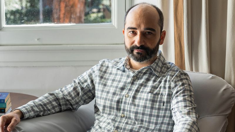 Color portrait of an olive skinned man in a button up shirt sitting in an armchair and looking at the camera