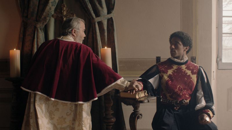 Color film still showing two men (one with dark skin, the other with light skinned) dressed in Renaissance era clothes in a room