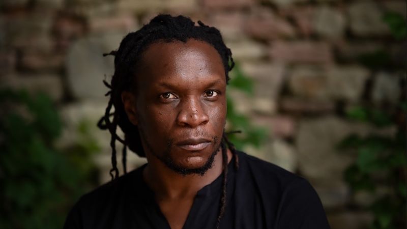 Color photograph of the head and shoulders of a dark skinned man with locks tied behind his head, looking at the camera with a serious expression
