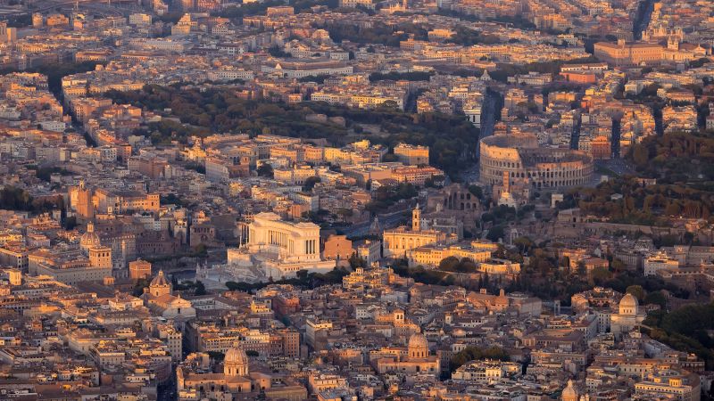 Color photograph of an aerial view of Rome taken from a helicopter, showing a broad cityscape bathed in rose colored light
