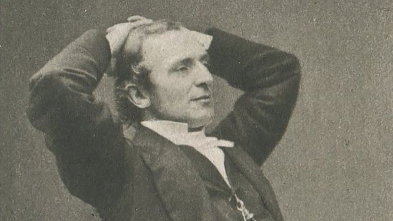 19th century photograph of a light skinned man sitting in a chair in a 3/4 pose; his arms are raised with his hands touching the back of his head