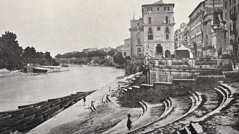 Monochrome photograph from the late 19th century showing a river on the left and a few scattered buildings on the right