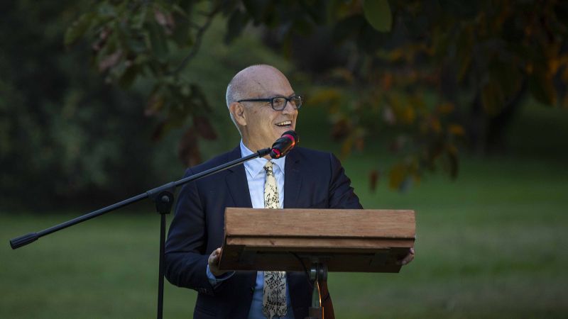 Color photograph of an older light skinned man standing in an outdoor garden at a podium and speaking into a microphone