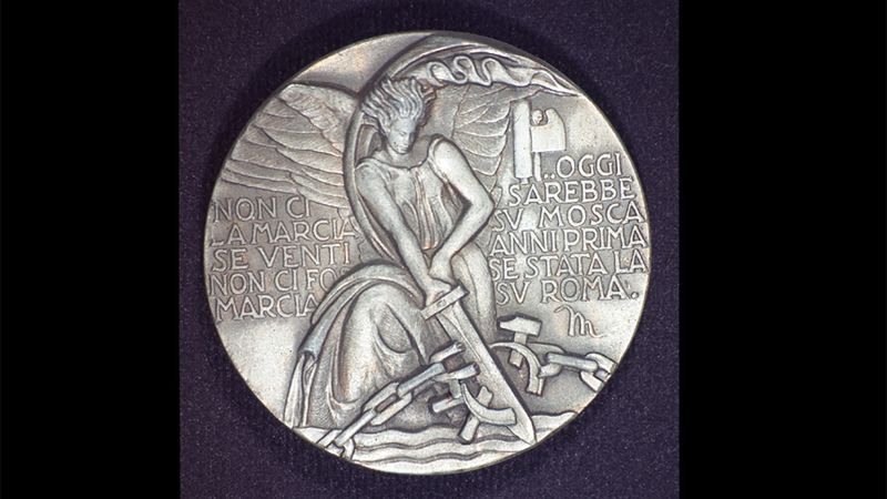 Silver colored round medallion with text and a winged woman breaking a chain with a sword
