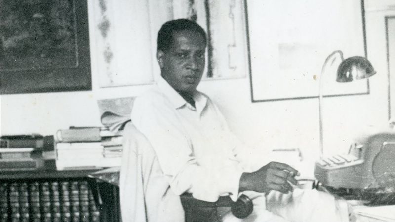 Black and white photograph of William Demby sitting in a chair at his desk in front of a typewriter, with framed art on the walls and a bookshelf visible behind him