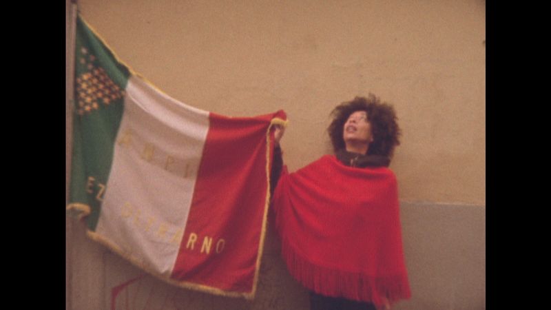 Color film still of a dark skinned woman wearing red and holding the edge of the tricolored Italian flag