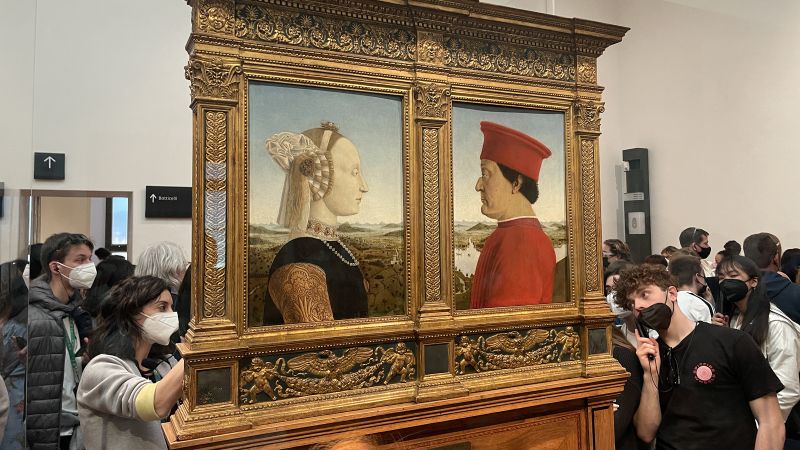 color photograph of a room inside the Uffizi Gallery with two profile painted portraits in a freestanding gold colored frame; surrounding the work is a crowd of people wearing facemasks and listening to audio guides