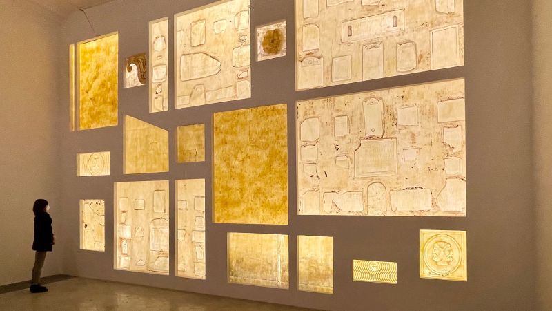 Color photograph of eighteen latex imprints of outdoor walls, some bearing ancient latin inscriptions, at the American Academy in Rome, hung in an interior gallery space; lighting is low but the yellow and golds of the latex are prominent; a woman stands to the far left to give scale