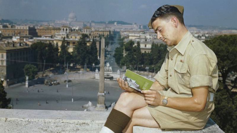 Color photograph of a man in profile, dressed in khaki shirt and shorts, sitting in an urban outdoor location overlooking a Roman plaza and reading a guidebook of Rome