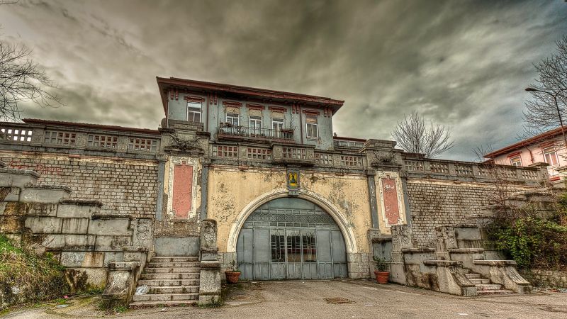 Color photograph of the facade of an abandoned, crumbling early-twentieth-century hospital administration building, with an overcast sky