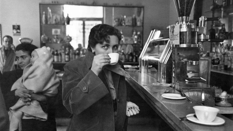 Black and white photograph of a stylish modern woman drinking an espresso at a bar; behind her is a young Italian mother holding a small child
