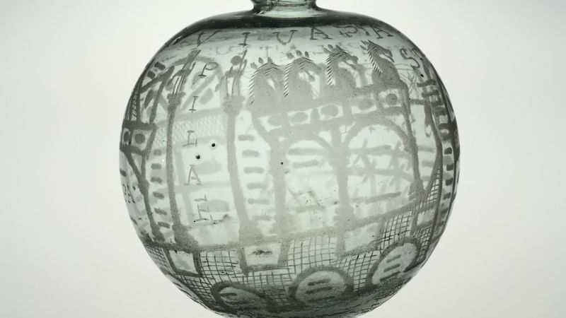 Color photograph of a very pale green ancient glass vessel painted with architectural elements, horses, and geometric patterns