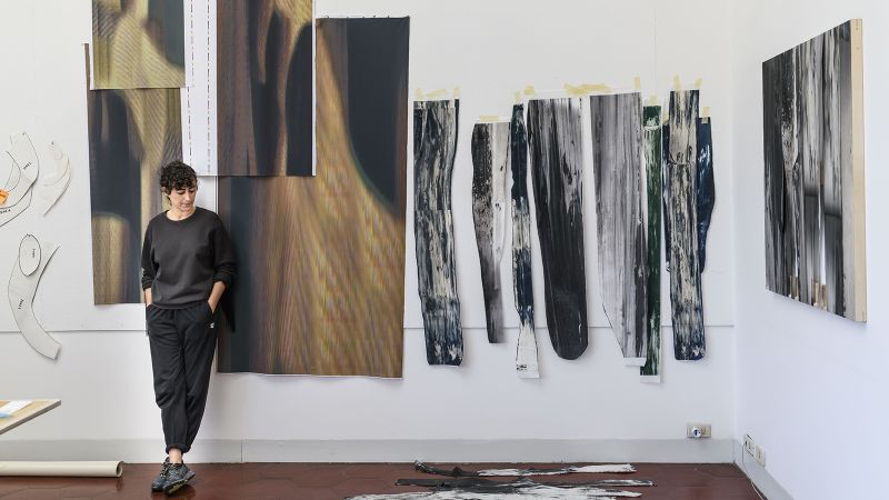 Color photograph of Sara Enrico standing in her studio next to her artwork, hanging on the walls beside her