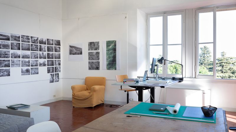 Color photograph of a view of 2022 Fellow Phoebe Lickwar's studio, showing several dozen black and white photos arranged in a grid on two walls, a soft cushioned chair, a desk with a laptop and lamp, and large windows letting the sun in