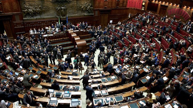 Balcony view of the interior of Italian parliament with politicians milling about a semicircle of desks and chairs
