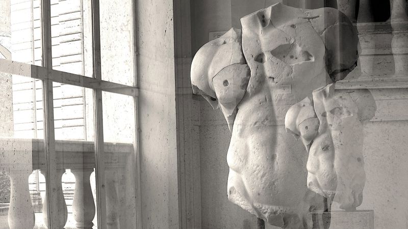 Black and white photograph of the torso of an ancient Roman sculpture (without a head, legs, and arms) next to a paned glass window