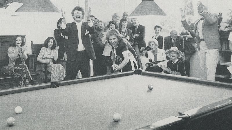 Black and white photograph from 1994 of many light skinned people clapping and cheering on a woman shooting pool in a salon