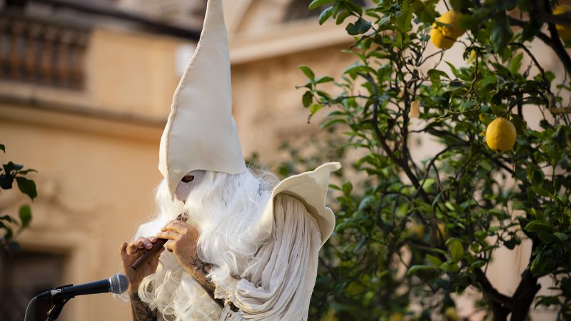 Color photograph of a man wearing a white costume and hood, standing in front of potted lemon trees, and playing a flute like instrument into a microphone