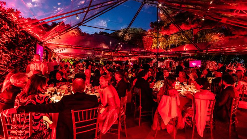 Color photograph of dozens of people sitting at round tables at a benefit gala; the room is lit with red lights