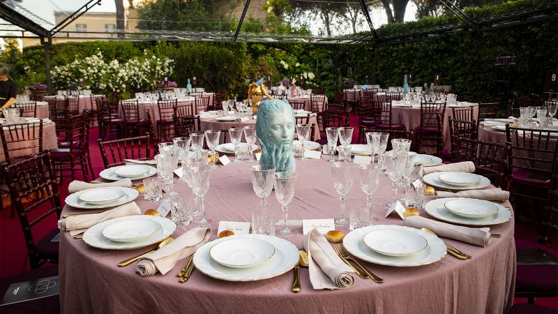 Color photograph of a round dinner table set with plates and silverware; in the middle is a green sculpture of a human head