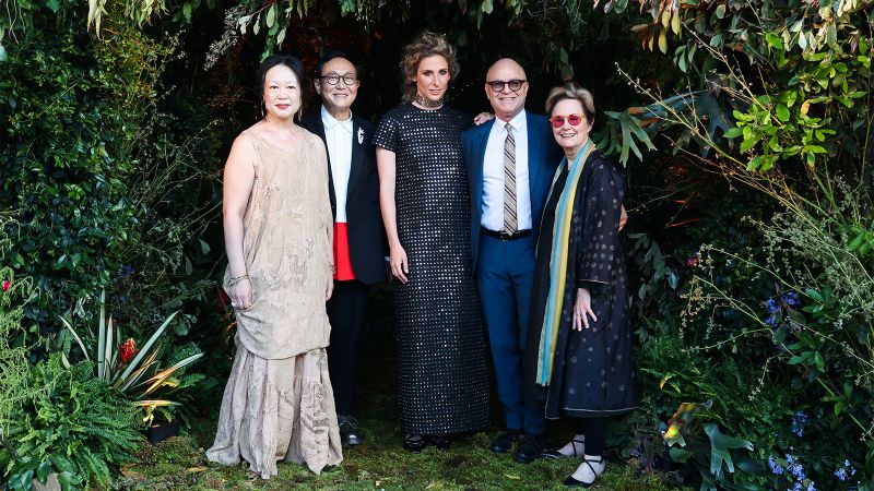 Color photograph of two women and three men wearing fancy gala clothes and standing in a single row in front of a thick garden background