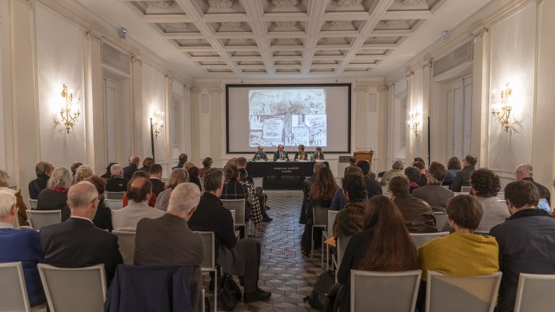 Color photograph showing a conference session in progress, with audience members, a panel of speakers, and a projection screen